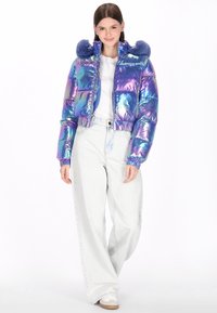 Prześwitująca krótka kurtka puchowa z niebieskim futrzanym kapturem, elastycznymi mankietami i błyszczącym wykończeniem, w połączeniu z jasnymi dżinsami i białymi tenisówkami.