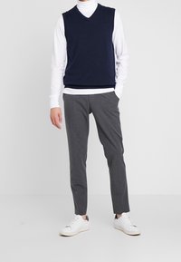 Marineblauer ärmelloser Pullover über einem weißen Rollkragenpullover, kombiniert mit grauen Hosen und weißen Turnschuhen. Schlichtes Design mit V-Ausschnitt.