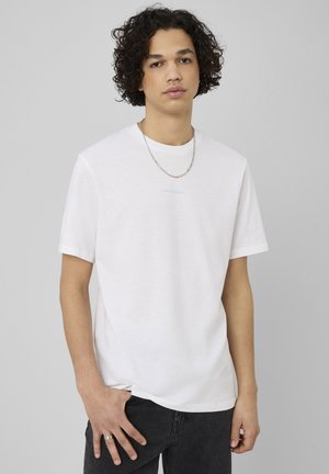 Jeune homme aux cheveux bouclés portant un t-shirt blanc uni, une chaîne en argent, un jean noir et une bague à la main gauche.