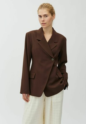 Femme aux cheveux blonds portant un blazer marron foncé à un seul bouton et un pantalon blanc ample texturé, debout devant un fond clair uni.