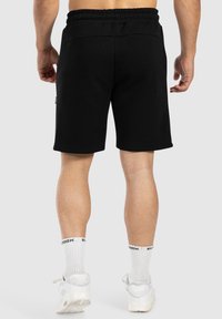 Pantalones cortos de algodón negros con una cinturilla elástica, que llegan a la mitad del muslo, con bolsillos laterales y una textura suave. Usados con calcetines y zapatos blancos.
