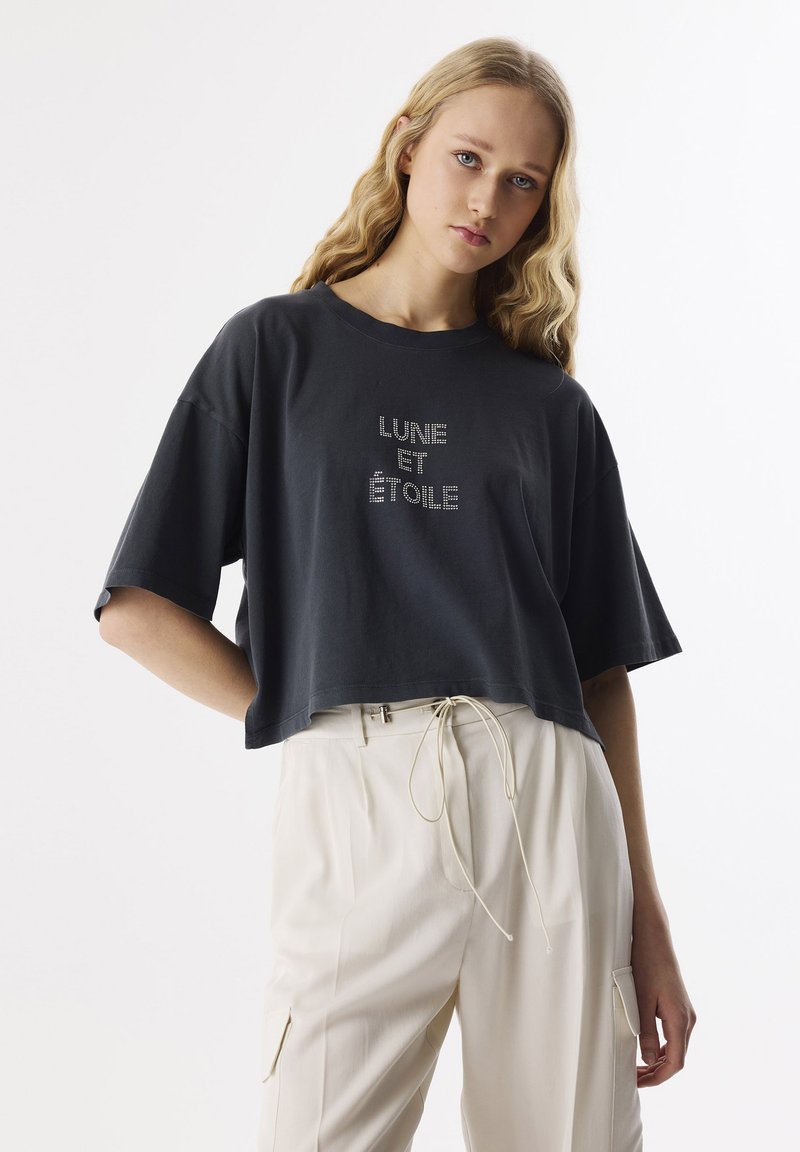 Camiseta negra recortada con texto en strass plateado "LUNE ET ÉTOILE"; combinada con pantalones beiges que cuentan con cordón y bolsillos laterales.