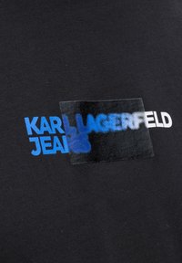 Juodas audinys su tekstūros raštu. Atrodo, kad spausdinta tekstas: "KARL" mėlyna spalva, "JEANS" šviesiai mėlyna, su skaidriu juodu bloku virš "LAGERFELD."