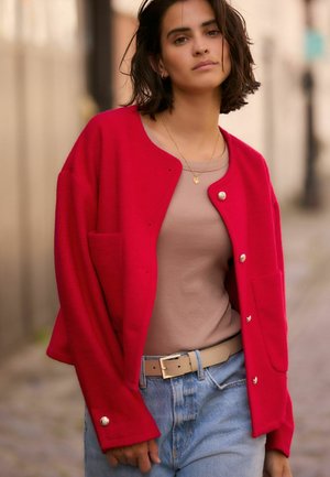 Veste en laine rouge avec des boutons en argent et des poches avant, portée sur un haut beige et un denim bleu clair. Texture douce et coupe décontractée.