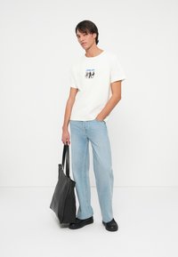 Młody mężczyzna w białej koszulce z grafiką i jasnoniebieskich dżinsach, trzymający dużą czarną torbę na ramię, stojący na tle białej ściany.