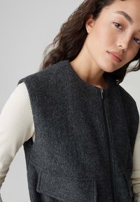 Gilet grigio in feltro senza maniche con collo alto e zip frontale, dotato di due tasche frontali e indossato sopra una maglia bianca a maniche lunghe a coste.