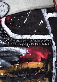 Denim noir avec des éclaboussures de peinture abstraites en rouge, blanc et or. Présente une bande à clous noire le long d'une poche et des bords effilochés.
