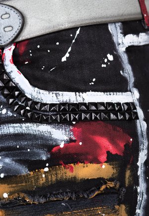 Denim noir avec des éclaboussures de peinture abstraites en rouge, blanc et or. Présente une bande à clous noire le long d'une poche et des bords effilochés.