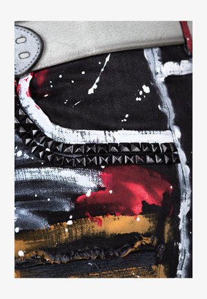 Denim noir avec des éclaboussures de peinture abstraites en rouge, blanc et or. Présente une bande à clous noire le long d'une poche et des bords effilochés.