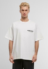 White cotton T-shirt with oversized fit, round neckline, short sleeves, and printed text “ANOTHER COTTON WORLDWIDE” on left chest.