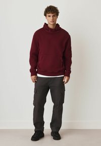 Bordeaux hoodie met een voorzak, over een wit shirt, gecombineerd met donkergekleurde cargobroek met zijzakken en zwarte sneakers.