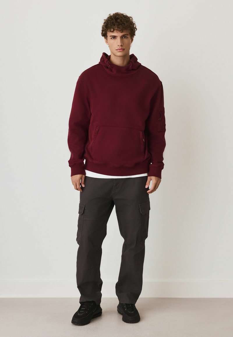 Bordeaux hoodie met een voorzak, over een wit shirt, gecombineerd met donkergekleurde cargobroek met zijzakken en zwarte sneakers.