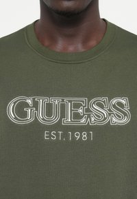 Olívovo zelené bavlnené tričko s rebrovaným okrúhlym výstrihom, s bielym vyšívaným logom "GUESS" a nápisom "EST. 1981" pod ním.