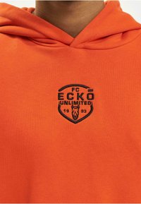 Oransje hettegenser i bomull med en svart brodert logo med hestedesign, "FC ECKÖ UNLIMITED" og "1993" under.