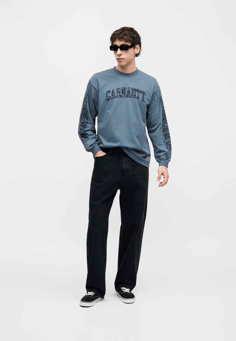 Junger Mann mit schwarzen Sonnenbrillen, blauem Carhartt-Langarmhemd, schwarzen Hosen und schwarzen Sneakers, stehend vor einem schlichten weißen Hintergrund.