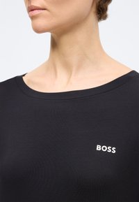 Γυναίκα που φοράει μαύρο πουκάμισο με στρογγυλή λαιμόκοψη και μικρό λευκό λογότυπο "BOSS" στο στήθος, εμφανιζόμενη από τον λαιμό έως το άνω μέρος του κορμού.