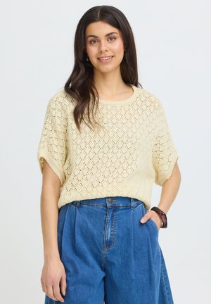 Młoda kobieta o długich ciemnych włosach, ubraną w beżowy, krótki sweter z krótkim rękawem i niebieskie dżinsy z wysokim stanem, uśmiechająca się z jedną dłonią w kieszeni.