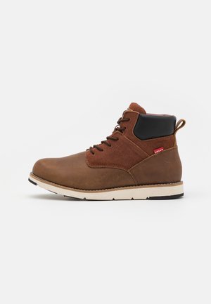 Botas de tobillo de cuero marrón con parte superior de ante, cuello sintético negro, entresuela blanca y suela de goma negra. Cuenta con etiqueta de logo roja.