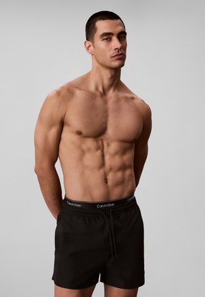 Hombre musculoso con cabello corto que lleva pantalones cortos negros de Calvin Klein, de pie contra un fondo gris liso con las manos detrás de la espalda.