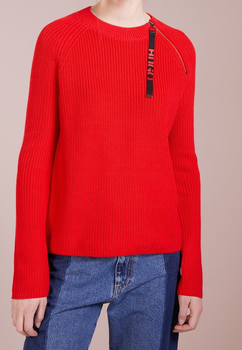 Pull rouge en maille texturée côtelée, col rond et fermeture éclair sur l'épaule. Associé à un jean bleu.