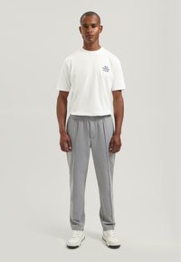 Bărbat în picioare, purtând un tricou alb cu mesajul "No Strezz Club", pantaloni gri cu cusături și adidași albi, pe un fundal simplu și luminos.