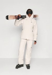 Άτομο σε κρεμ σκι στήσιμο και μαύρες μπότες, κρατώντας σκι πάνω από τον ώμο, φορώντας μαύρα γυαλιά σκι σε απλό φόντο.