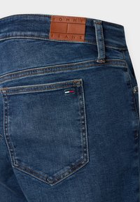 Σκούρο μπλε τζιν υφασμένο από ντένιμ με ματ υφή, διαθέτοντας ένα καφέ δερμάτινο ταμπελάκι με ανάγλυφο το λογότυπο "TOMMY JEANS" στη μέση και μικρή διακριτική λεπτομέρεια λογότυπου στην τσέπη.