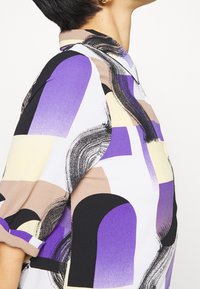 Personne portant une chemise à col avec des manches retroussées ornée d'un motif abstrait en violet, noir, beige et blanc.