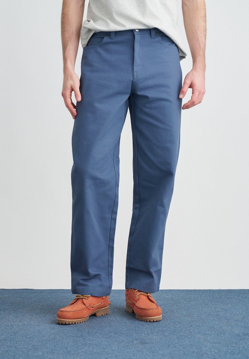 Μπλε παντελόνι από ύφασμα με ίσια γραμμή, χαρακτηρίζεται από εμπρόσθιες τσέπες και διακριτικές ραφές. Συνδυάζεται με πορτοκαλί παπούτσια.