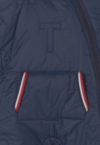 Tummansininen tikkitakki, jossa on tekstuurilla varustettu "T"-logo ja vetoketjullinen tasku punaisilla, valkoisilla ja sinisillä korostusraidoilla. Pehmeä ja kevyt materiaali.