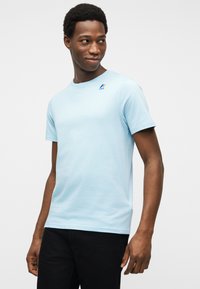 Νεαρός άνδρας που φοράει ανοιχτό μπλε κοντομάνικη μπλούζα με μικρό λογότυπο στο στήθος, όρθιος και κοιτάζοντας πλάγια σε απλό φόντο.