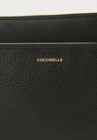 Clutch in pelle nera con superficie testurizzata, chiusura con zip dorata e logo "Coccinelle" in rilievo dorato.