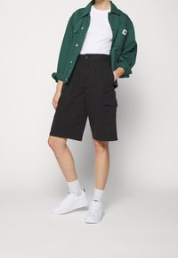 Shorts cargo noirs avec poches latérales, associés à un t-shirt blanc et une surchemise verte, complétés par des baskets blanches et des chaussettes.