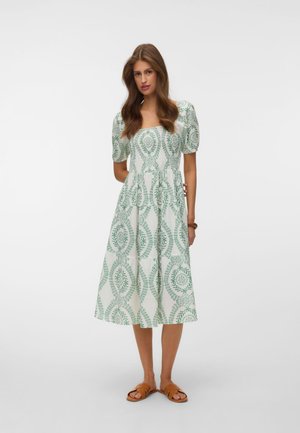 Kvinde iført en hvid midi-kjole med grønne blomsterprint, pufærmer og brune slide-sandaler, stående mod en ensfarvet hvid baggrund.