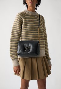 Saco de ombro em couro preto com alça de corrente dourada, apresentando um logótipo decorativo na parte da frente. Usado por cima de um suéter riscado e uma saia plissada.