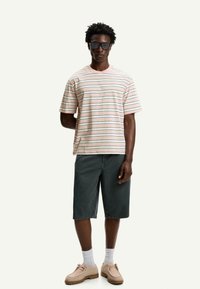 Striped cotton t-shirt in pastel colors, paired with dark denim shorts. Model wears beige shoes and black sunglasses. Neutral background.
