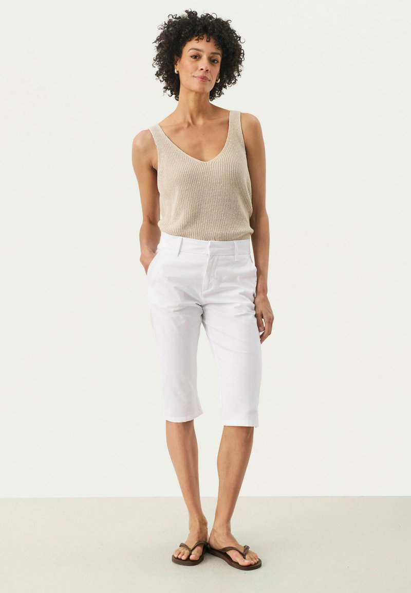 Débardeur en tricot beige avec un col en V, associé à un pantalon blanc raccourci et des tongs marron. Design simple et ajusté sans motifs.