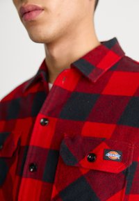 Chemise en flanelle à carreaux rouges et noirs avec col, poches à boutons et patch logo. Le matériau semble doux et texturé.
