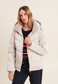Doudoune beige clair avec une capuche doublée en fausse fourrure, design matelassé, fermeture éclair à l'avant et poches latérales. Portée par-dessus un pull à rayures sombres.