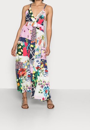 Kobieta ubrana w kolorową suknię maxi w stylu patchwork z cienkimi ramiączkami i kwiatowymi, owocowymi oraz abstrakcyjnymi wzorami, połączoną z brązowymi sandałami z paskiem wokół kostki.