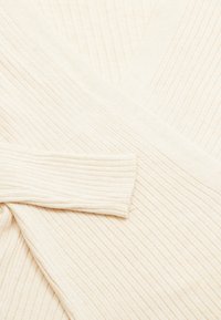 Pulover crem cu caneluri, cu un design în formă de V, având un material texturat și mâneci lungi. Materialul moale evidențiază o siluetă structurată.