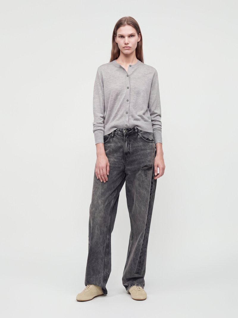 Femme portant un cardigan gris boutonné, un jean noir ample délavé et des chaussures beiges, debout devant un fond blanc uni.