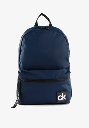 Sac à dos bleu marine Calvin Klein avec fermeture éclair noire, poignée supérieure, poche avant et écusson logo blanc "ck" en bas à droite à l'avant.