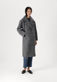 Cappotto doppiopetto grigio in lana con ampio collo a risvolto, tasche laterali e vestibilità rilassata, abbinato a jeans a vita bassa e scarpe nere slip-on.
