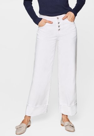 Personne portant un pantalon blanc à jambes larges avec braguette à boutons, un haut bleu marine à manches longues et des chaussures beige à enfiler avec un détail nœud, mains dans les poches.