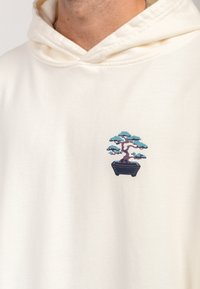 Grietos spalvos džemperis su mažais siuvinėtais bonsai medžių dizainu žalios ir violetinės spalvų atspalviuose, su juodu vazonu, išsamiai parodytu apačioje.