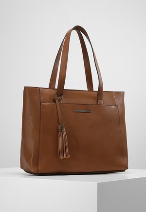 Sac fourre-tout en cuir marron avec des bretelles doubles et un détail de pompon sur la poche avant, présenté sur un socle blanc contre un fond neutre.