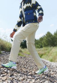 Virkningsjacka med blå och grön mönster, beige cargobyxor och flerfärgade sneakers, som går på en stenig stig med grönska i bakgrunden.