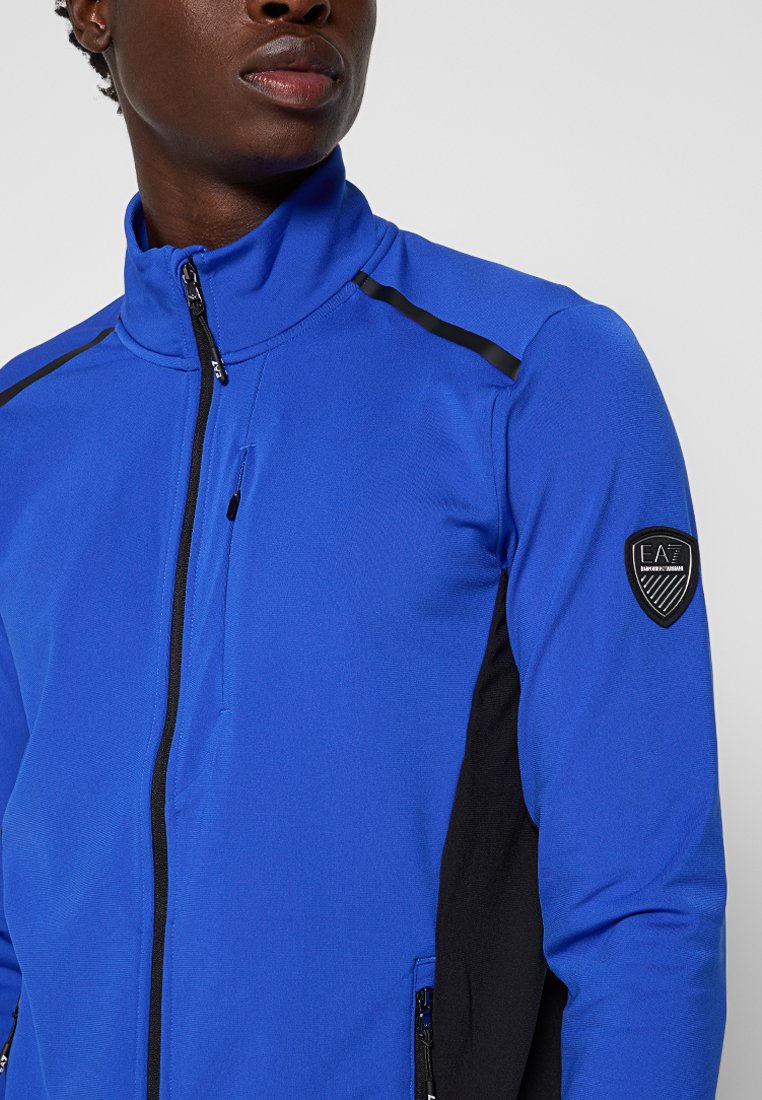 Giacca blu con zip, colletto alto, dettagli neri alle spalle e ai lati, tessuto texture e una patch del logo sulla manica sinistra.
