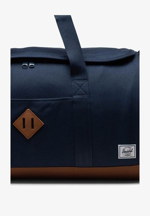 Navy fabric duffel bag with brown leather accents, featuring a rectangular shape, top zip closure, and a small brand label.